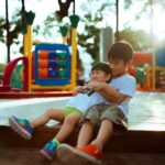 Zwei Kinder auf einem Spielplatz. Ein älterer Junge umarmt ein jüngeres Mädchen, während sie lachend auf einem Holzpodest sitzen, umgeben von bunten Spielgeräten